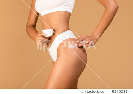 Unrecognizable slim lady in white underwear applying nourishing body lotion on her hips, beige background 93202314