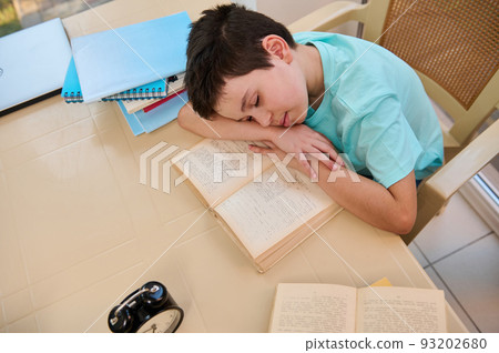 Top view of a tired sleepy schoolboy falling asleep on a textbook while doing homework. Homeschooling. Daily routine of a student. Learning disability. Difficulties in restoring the school regime Top view of a tired sleepy schoolboy falling asleep on a textbook while doing homework. Homeschooling. Daily routine of a student. Learning disability. Difficulties in restoring the school regime 93202680
