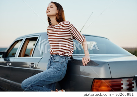 A young woman takes a break from driving, leaning on her car beside the road and smiling and looking at nature and the sunset A young woman takes a break from driving, leaning on her car beside the road and smiling and looking at nature and the sunset 93203991