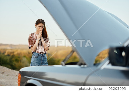 Woman traveler standing near the car with the hood open and looking for the cause of the car breakdown alone without men on the road in the countryside 93203993