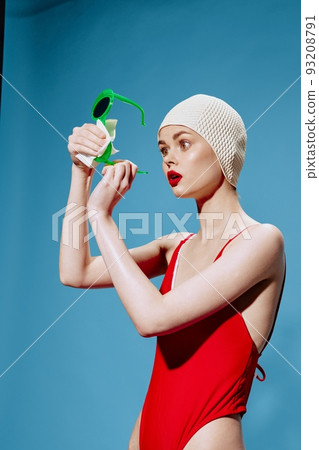 An enthusiastic shocked unusual girl in a swimming cap and a red swimsuit wipes glasses in the studio on a blue background 93208791