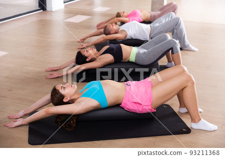 Group of people performs an exercise by placing a roll of a sports mat under their back Group of people performs an exercise by placing a roll of a sports mat under their back 93211368