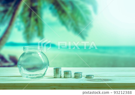 Financial concept, Empty glass bottle to show the money spent on vacation or holidays and stack coins to show decrease of money on wooden table, coconut tree and beach blurred background. 93212388