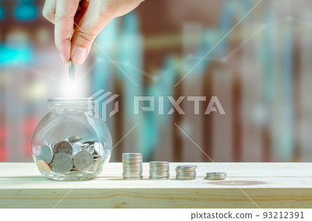 Saving money and investment concept, hand putting coin in glass bottle for savings and stack coins to show increase of savings money on wooden table, graphs and index blurred background. 93212391