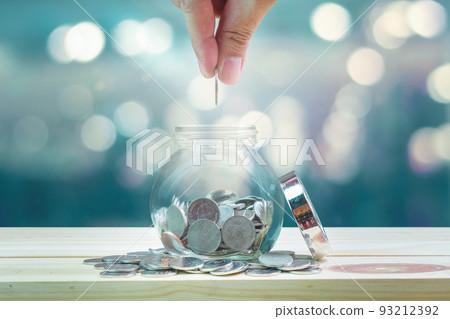 Saving money and investment concept, hand putting coin in glass bottle for savings which on wooden table and bokeh background. 93212392