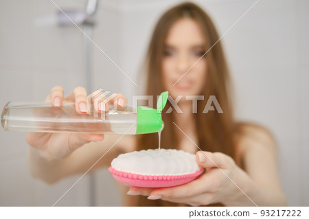 Defocused woman stands in bathroom cabin squeezing refreshing shower gel on a washcloth 93217222