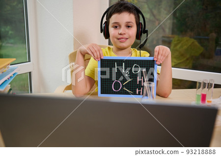 Smart schoolboy in wireless headphones, shows a blackboard with a chemical formula to the teacher during an online chemistry lesson. Homeschooling. Back to school. New semester of the academic year Smart schoolboy in wireless headphones, shows a blackboard with a chemical formula to the teacher during an online chemistry lesson. Homeschooling. Back to school. New semester of the academic year 93218888
