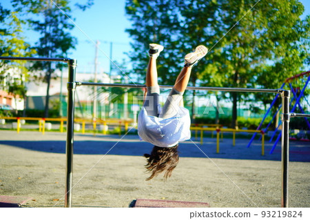 A girl practicing inversions on a horizontal bar in the park. Summer vacation of the 3rd grade of elementary school. A girl practicing inversions on a horizontal bar in the park. Summer vacation of the 3rd grade of elementary school. 93219824