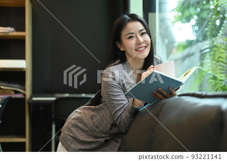 Cheerful asian working woman relaxing on sofa in her bright modern office and reading book Cheerful asian working woman relaxing on sofa in her bright modern office and reading book 93221141