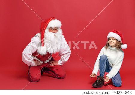 Cute little girl in Christmas hat trying on a boot with Santa against red studio background. Isolate Cute little girl in Christmas hat trying on a boot with Santa against red studio background. Isolate 93221848