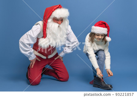Cute little girl in Christmas hat trying on a boot with Santa against blue studio background. Isolate Cute little girl in Christmas hat trying on a boot with Santa against blue studio background. Isolate 93221857