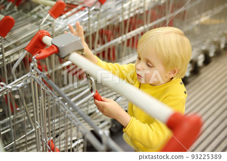 A cute blonde boy plays with shopping carts at the entrance to the supermarket A cute blonde boy plays with shopping carts at the entrance to the supermarket 93225389