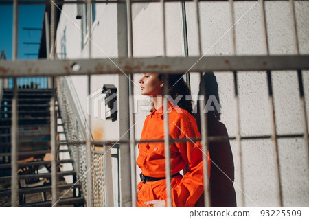 Young brunette curly woman in orange suit behind cage. Female in colorful overalls portrait Young brunette curly woman in orange suit behind cage. Female in colorful overalls portrait 93225509
