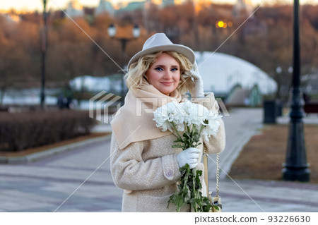 A blonde woman in a coat, hat and gloves with a bouquet of white chrysanthemums and a cup of coffee on the background of an arch and an autumn landscape with a road on an autumn evening at sunset 93226630
