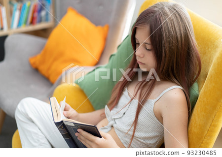A young girl is reading a book while sitting in a bright yellow armchair in a public library. The concept of home education, recreation, mindfulness. The girl is wearing light clothes , side view . 93230485