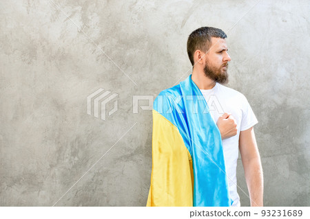 Man holds hand over his heart and national blue-yellow flag of Ukraine on his shoulder. Guy in white T-shirt and jeans. Sunny day. Constitution and Independence Day of Ukraine. Soft selective focus Man holds hand over his heart and national blue-yellow flag of Ukraine on his shoulder. Guy in white T-shirt and jeans. Sunny day. Constitution and Independence Day of Ukraine. Soft selective focus 93231689