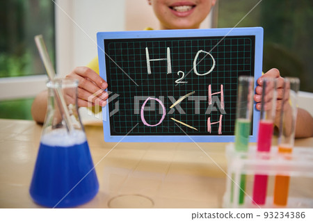 Focus on blackboard with the chemical formula of water written with colored chalk in hands of smiling little chemist sitting at table with test tubes on tripod and flask with chemical liquid, reagents 93234386