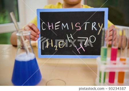 Blackboard with written inscription Chemistry, made with chalk in the hands of a schoolboy chemist, sitting at a desk with flask and test tubes filled with chemical liquids and reagents on the tripod 93234387