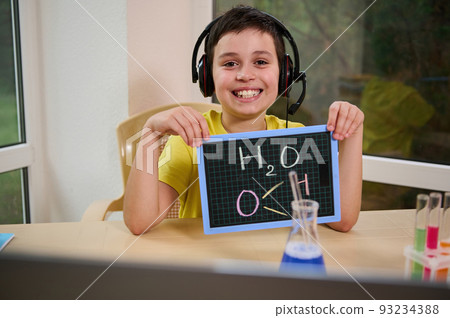 Happy schoolboy wearing audio headset, smiles a toothy smile, sitting at laptop and showing a blackboard with the chemical formula of water H2O to the teacher during online chemistry lesson 93234388