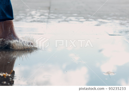 Woman wearing rain rubber boots walking running and jumping into puddle with water splash and drops in autumn rain. 93235233