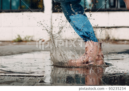 Woman wearing rain rubber boots walking running and jumping into puddle with water splash and drops in autumn rain. Woman wearing rain rubber boots walking running and jumping into puddle with water splash and drops in autumn rain. 93235234