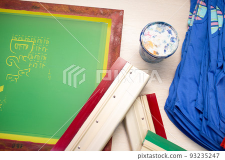 Serigraphy silk screen print process at clothes factory. Frame template stencil, squeegee and plastisol paint at table with cloth. Serigraphy silk screen print process at clothes factory. Frame template stencil, squeegee and plastisol paint at table with cloth. 93235247