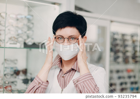 Happy Optician in Medical Face Mask Choosing Eyeglasses, female optometrist with glasses. 93235794