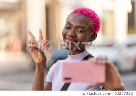 Smiling african woman taking selfie while gesturing victory outdoors 93237330