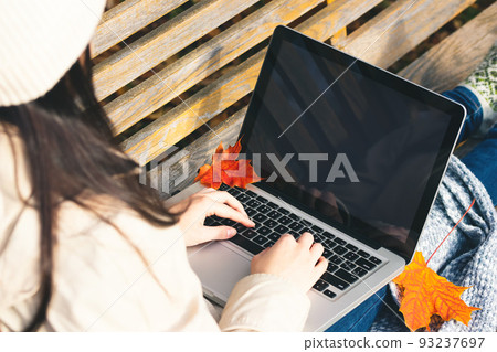 Girl uses a laptop while sitting on a bench in the autumn park. Blank for design on laptop screen 93237697
