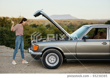 A woman opens the hood of a broken down car and tries to find the cause of the breakdown on the road during a nature trip alone and is sad 93238385