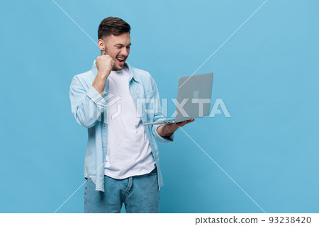 Happy lucky tanned handsome man in casual basic t-shirt raises fist up gesture success with laptop posing isolated on blue studio background. Copy space Banner Mockup. Electronics repair IT concept 93238420