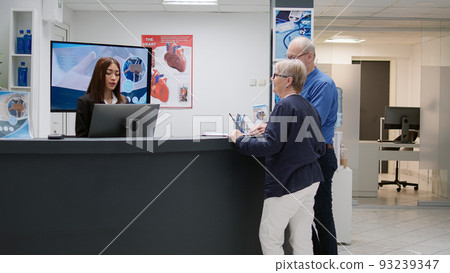 Retired people doing medical report with receptionist in hospital waiting room, woman at reception desk helping patients with checkup appointment. Assistant working in healthcare center. Retired people doing medical report with receptionist in hospital waiting room, woman at reception desk helping patients with checkup appointment. Assistant working in healthcare center. 93239347