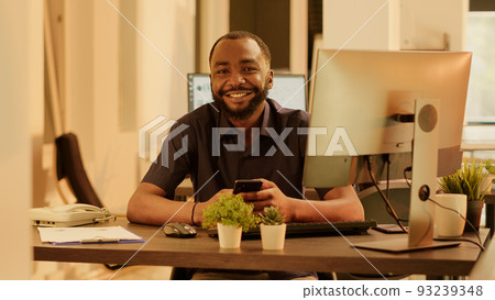 Portrait of african american man using smartphone app on computer, planning business presentation and doing research on social media network. Working with internet application on mobile phone. 93239348