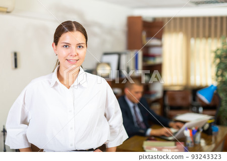 Portrait of a smiling young female employee standing in a office Portrait of a smiling young female employee standing in a office 93243323