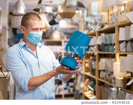 Man in protective mask is choosing new table lamp for her home in store Man in protective mask is choosing new table lamp for her home in store 93243610
