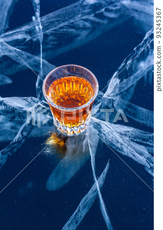 A glass of whiskey stands on beautiful ice with deep cracks. Orange liquid on a blue background. Top view from the side. Bizarre white deep cracks in the ice. Vertical. A glass of whiskey stands on beautiful ice with deep cracks. Orange liquid on a blue background. Top view from the side. Bizarre white deep cracks in the ice. Vertical. 93245367