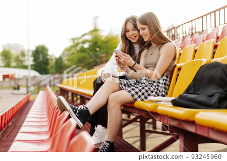 Two friends of a schoolgirl or a student are sitting in the stands and watching videos or communicating using a smartphone 93245960