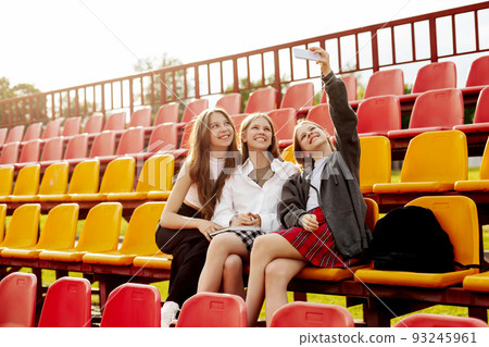 Three charming friends take selfies on their phone at the school stadium. The concept of friendship. Education, training, back to school Three charming friends take selfies on their phone at the school stadium. The concept of friendship. Education, training, back to school 93245961