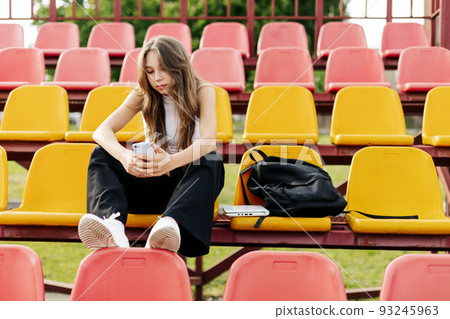 A teenage girl sits on the school bleachers and writes a message on her phone in her free time A teenage girl sits on the school bleachers and writes a message on her phone in her free time 93245963
