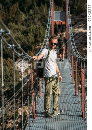 Traveler with a backpack in the mountains. A middle-aged man stands on a suspension bridge on a background of a forest in mountains. Adventure trip to Europe. 93248235