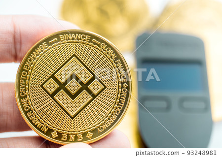 Hand holding a Binance Smart Chain Cryptocurrency BNB coin with hardware wallet in background. Hand holding a Binance Smart Chain Cryptocurrency BNB coin with hardware wallet in background. 93248981