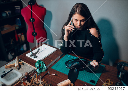 Tanner woman making leather harness and call client mobile phone on workshop. Working process of leather craftsman. 93252937