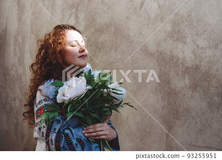 Girl with closed eyes holds bouquet of peonies. Woman in embroidered shirt and with long red curly hair against concrete wall. Photo dark key. Independence Day of Ukraine. Copy space. Soft focus 93255951