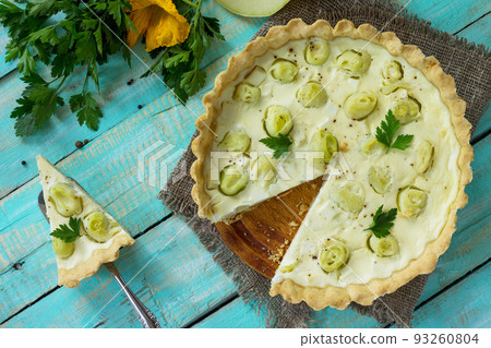 A classic quiche Lorraine pie with feta cheese with zucchini, egg pouring and cheese on a kitchen wooden table. Top view with copy space. 93260804