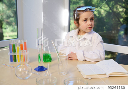 Adorable Caucasian child, little schoolgirl in protective eyeglasses and white lab coat, sitting at a table with laboratory glassware, enjoying fascinating chemical experiments in the chemistry lesson 93261180