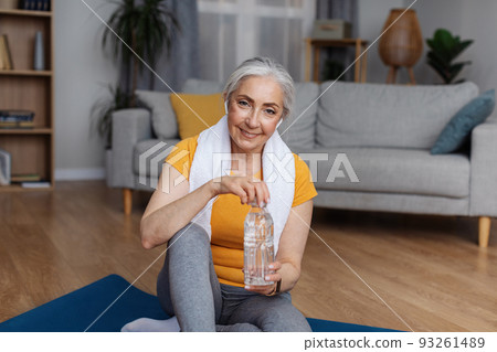 Senior woman in sportswear sitting on sports mat with bottle of clear water, keeping hydrated during domestic training Senior woman in sportswear sitting on sports mat with bottle of clear water, keeping hydrated during domestic training 93261489