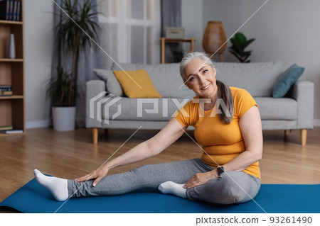 Home workout. Senior woman stretching her leg on yoga mat in living room and smiling at camera, copy space 93261490