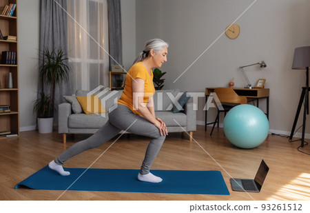 Sporty senior woman making lunges near laptop, working out to online sports tutorial at home in living room Sporty senior woman making lunges near laptop, working out to online sports tutorial at home in living room 93261512