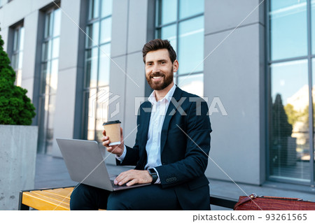 Smiling confident european millennial bearded businessman in suit work on laptop, drink coffee on bench 93261565