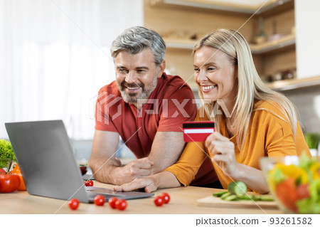 Cheerful couple making online order, using laptop at kitchen Cheerful couple making online order, using laptop at kitchen 93261582